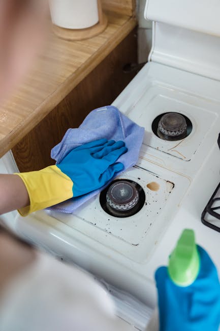 Close-up of a person wearing yellow and blue cleaning gloves using a damp cloth to wipe the burners of a white ceramic stovetop in a kitchen. The stovetop shows signs of grease and stains, indicating