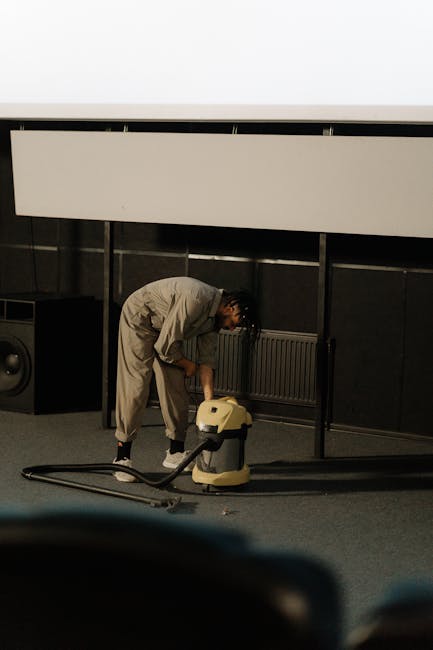 A person in a beige jumpsuit is performing deep cleaning on a dark carpeted floor of a room, operating a yellow industrial vacuum cleaner connected to a flexible hose. The room features a black wall w